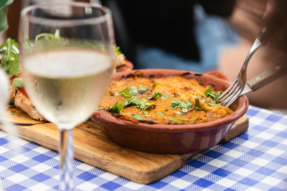 Tacho de barro gratinado e copo de vinho branco sobre toalha aos quadrados azuis